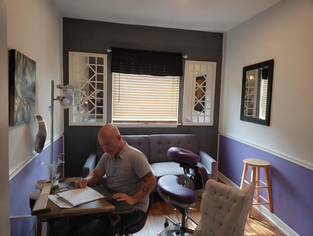 Bald man in a grey shirt with an arm tattoo sits at a wooden desk reading papers in a purple and grey room with a window