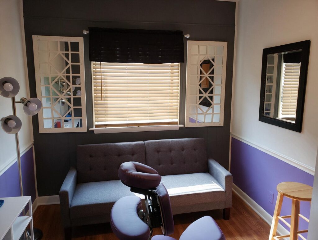Empty treatment room with a gray loveseat, purple massage chair, wood blinds, mirrors