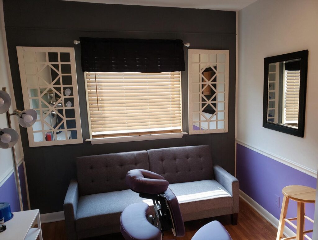 Empty treatment room with a gray loveseat under a blinded window, flanking white mirrors, purple chair