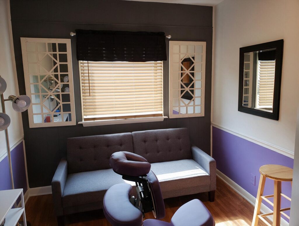 Empty room with a gray tufted loveseat under a blinded window, flanked by white lattice mirrors
