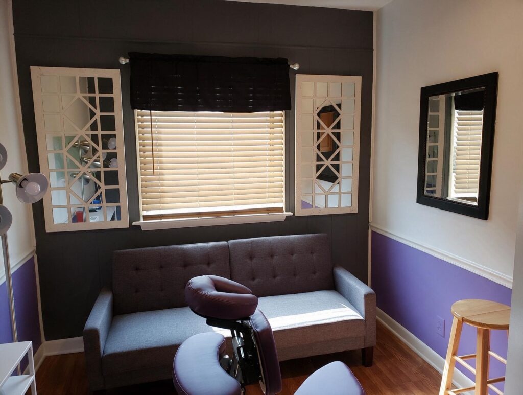 Empty treatment room with a grey sofa under a blinded window, two mirrors, purple and white walls