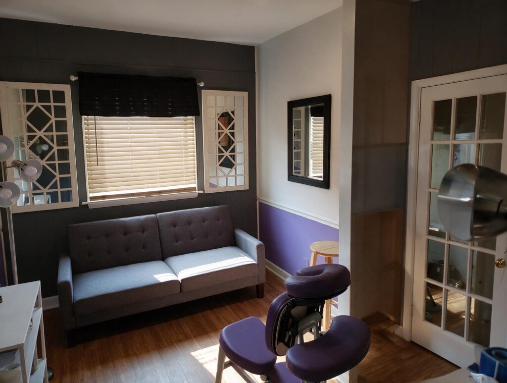 Empty treatment room with a gray sofa under a sunlit window, mirrors, a purple massage chair